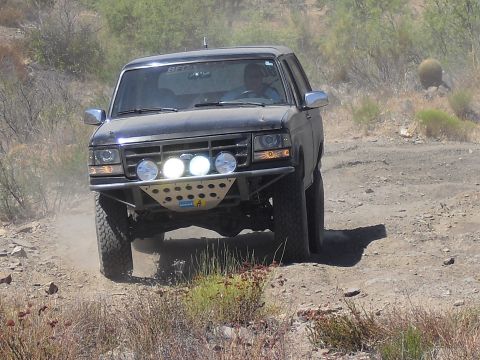 1986 Ford Bronco - Black Ford Bronco with off-road lights and bumper driving on a dirt road.