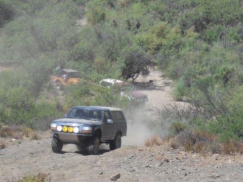 1986 Ford Bronco - Several off-road vehicles, including a Ford Bronco, navigate a dusty trail in a dry, scrubby landscape.
