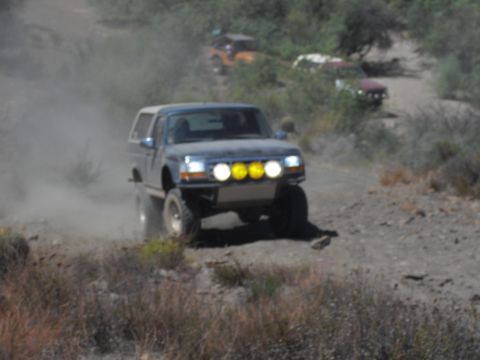 1986 Ford Bronco - A Ford Bronco kicks up dust while navigating a rough, off-road trail with other vehicles in the distance.