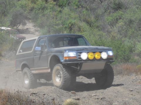 1986 Ford Bronco - A modified Ford Bronco navigates a dusty, off-road trail with other vehicles visible in the background.