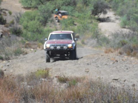 1986 Ford Bronco - Two off-road vehicles navigate a dusty trail in a dry, hilly landscape.