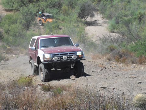 1986 Ford Bronco - Two off-road vehicles navigate a dusty trail through a dry, brush-covered hillside.