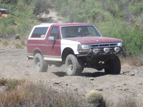 1986 Ford Bronco - A maroon and white Ford Bronco navigates a dusty, off-road trail with another vehicle visible in the background.