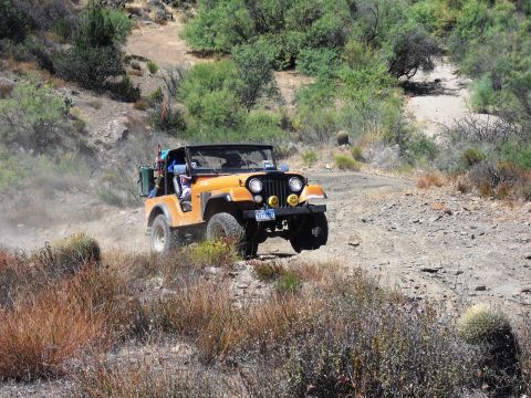 1986 Ford Bronco - An orange Jeep CJ-7 navigates a dusty, off-road trail surrounded by arid vegetation.