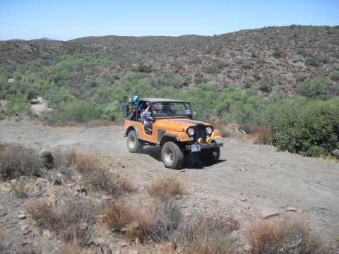 1986 Ford Bronco - An orange Jeep CJ-7 navigates a dusty, off-road trail through a dry, hilly landscape.