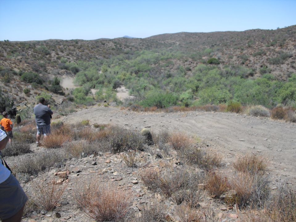 People stand on a rocky hillside overlooking a dry, brush-filled valley under a clear blue sky