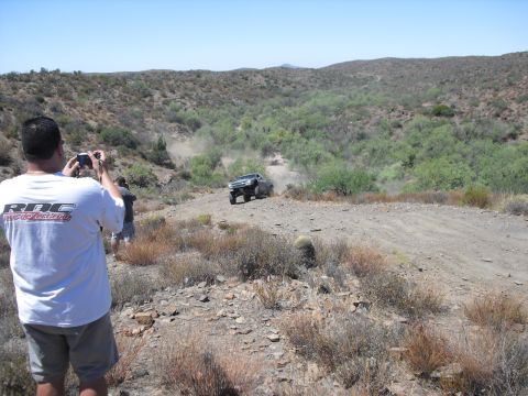 1986 Ford Bronco - A person photographs a vehicle navigating a dusty trail in a , arid landscape under a clear blue sky.