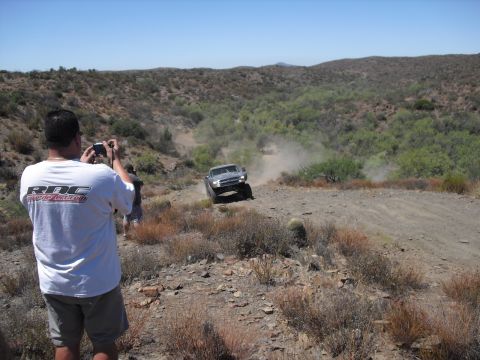 1986 Ford Bronco - A man photographs a silver SUV as it navigates a dusty off-road path in a dry, hilly landscape.