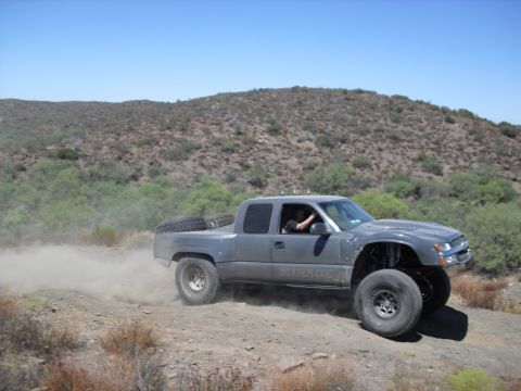 1986 Ford Bronco - A modified gray pickup truck navigates a dusty off-road trail under a clear blue sky.
