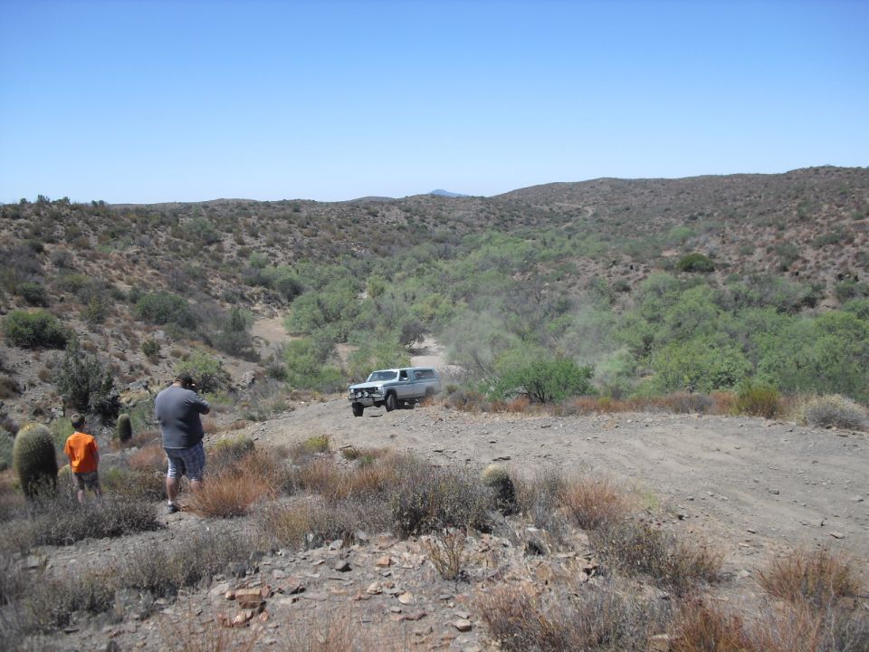 1986 Ford Bronco driving on a dusty dirt road in a desert with two people standing nearby