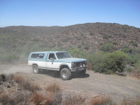 1986 Ford Bronco - This lifted pickup truck with a camper shell kicks up dust as it navigates a , off-road trail under a clear blue sky.