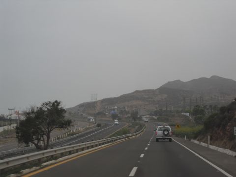 1986 Ford Bronco - A white SUV travels down a multi-lane highway under an overcast sky, with hills and distant structures visible.