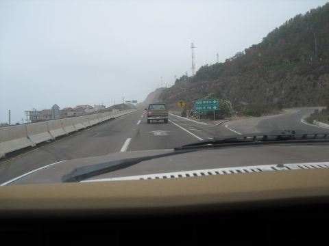 1986 Ford Bronco - A brown pickup truck travels ahead on a highway under a cloudy sky, with a mountainous landscape to the right.