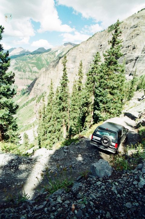 1995 Ford Bronco - A black SUV navigates a challenging mountain path, with a dramatic canyon and sky in the background.