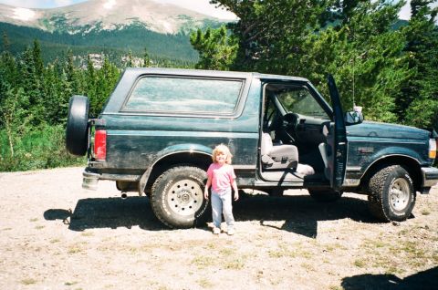 1995 Ford Bronco - A child stands beside a 1995 Ford Bronco parked in a mountainous, wooded area with the driver's door ajar.