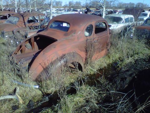 1978 Chevrolet C/K Pickup - A heavily rusted car sits abandoned in a junkyard, surrounded by other decaying vehicles.