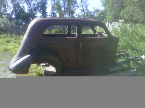 1978 Chevrolet C/K Pickup - This is a heavily rusted car body shell, viewed from the side, sitting on wooden planks in an outdoor setting.
