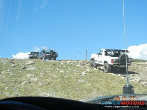 1995 Ford Bronco - Two off-road vehicles, a white 1995 Ford Bronco and a dark SUV, navigate a rocky incline under a clear blue sky.