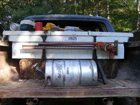 1978 Ford Bronco - The bed of a pickup truck holds a toolbox, a jack, a keg, and various other supplies, with trees in the background.