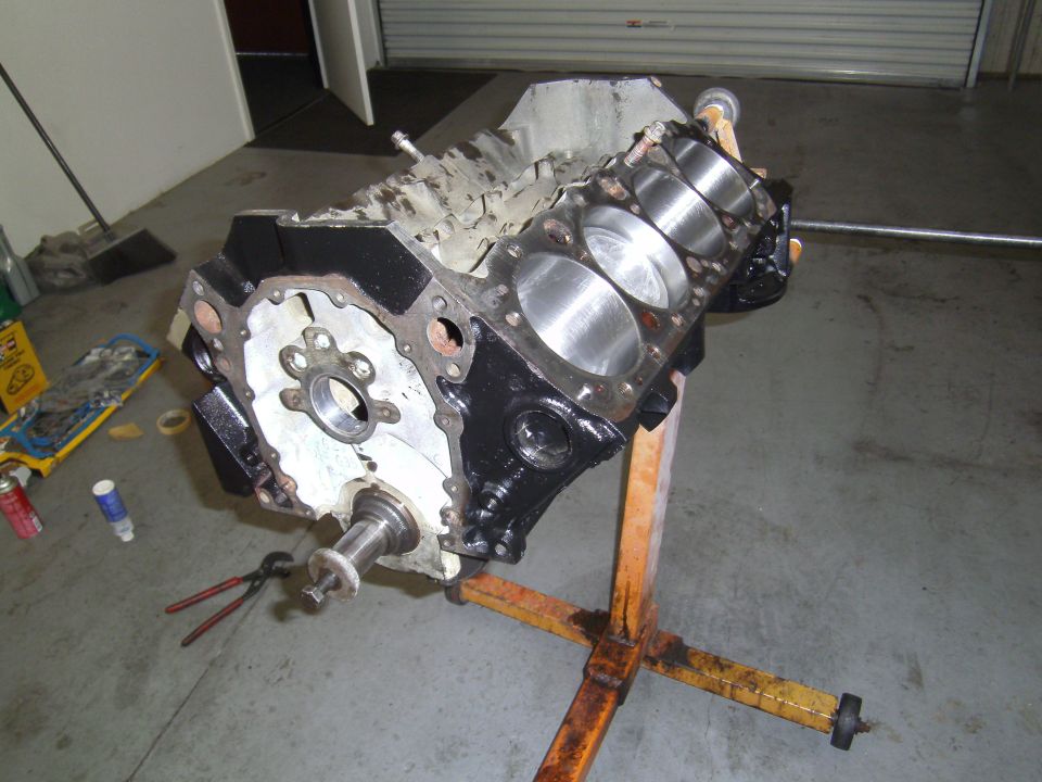 Engine block on an orange stand, showing cylinders and crankshaft flange. Pliers and spray cans are on the floor