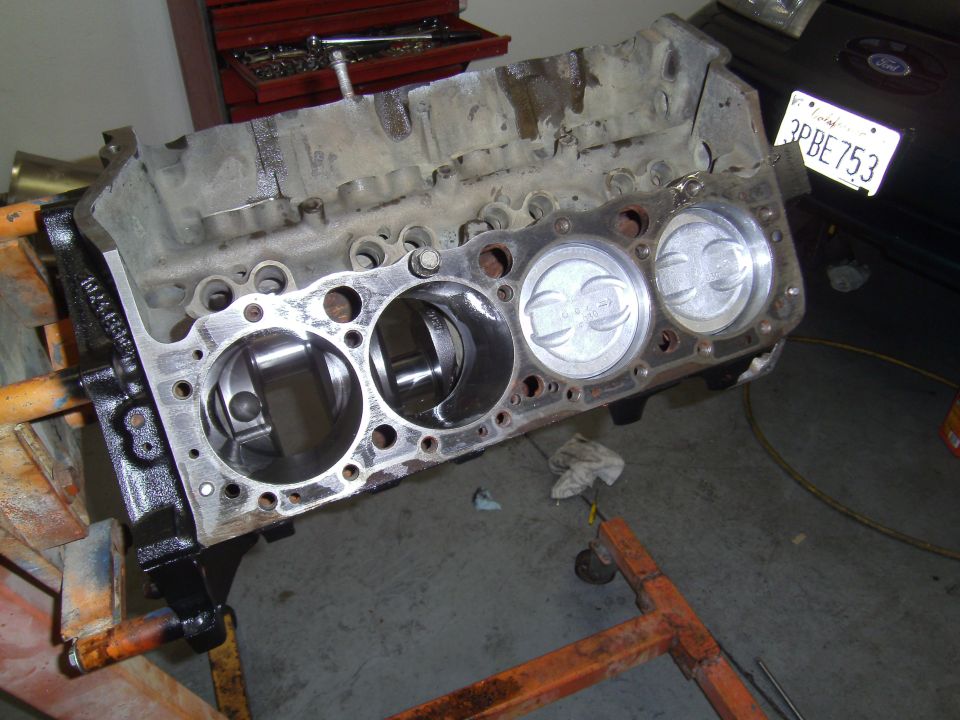 Engine block with three pistons visible, mounted on an orange engine stand. A license plate is partially visible in the background