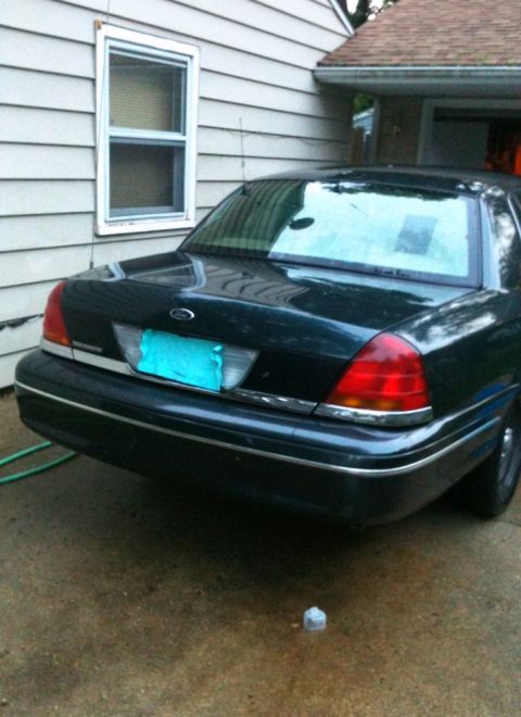 1998 Ford Crown Victoria - The rear of a dark 1998 Ford Crown Victoria is visible, parked on a wet driveway next to a house.
