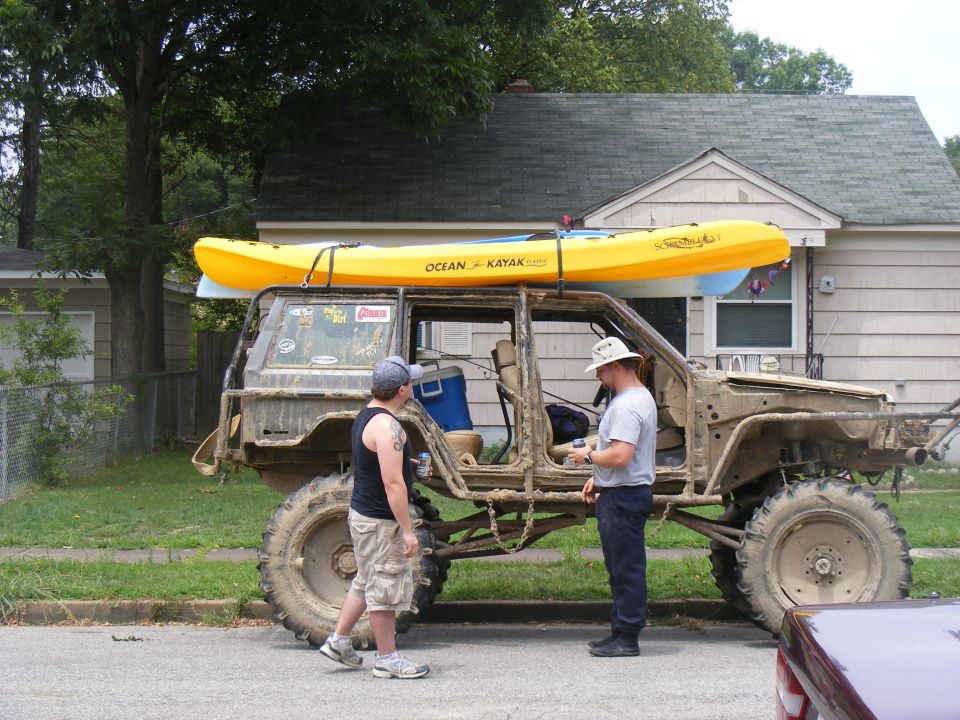 Mud-covered off-road vehicle with kayaks on roof rack, two men standing beside it