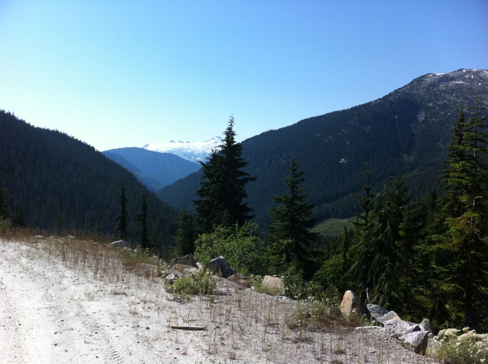 Mountain valley with snow-capped peaks, evergreen trees, and a dirt road in the foreground. Watermark reads SUPERMOTORS.NET