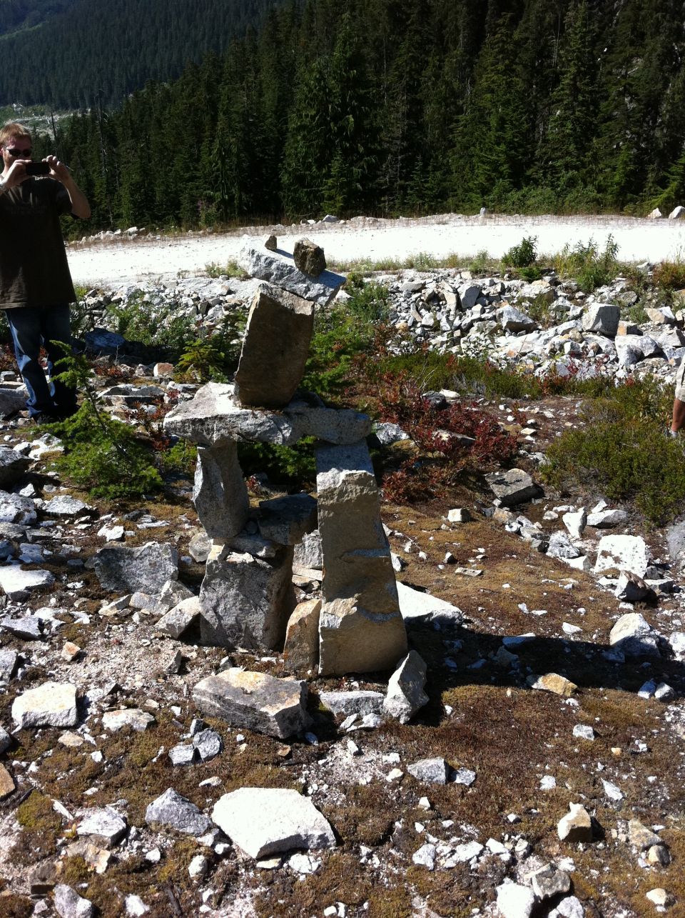 Person taking photo of stone inukshuk on rocky hillside with trees and dirt road in background
