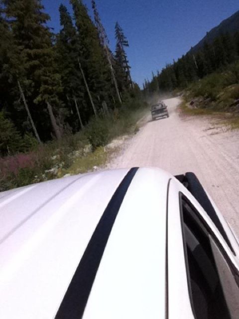 1990 Ford Bronco - Two trucks travel on a dusty dirt road through a wooded area.