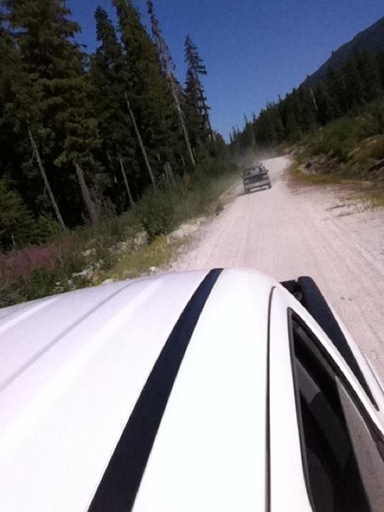 View from inside a white truck looking down a dusty dirt road with another truck ahead. Trees line the road