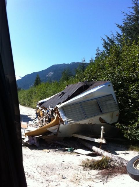 1990 Ford Bronco - A severely damaged travel trailer lies on its side on a dirt road, surrounded by debris and vegetation.