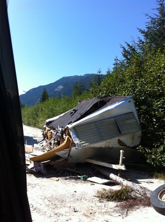 Wrecked travel trailer on a dirt road with trees and mountains in the background