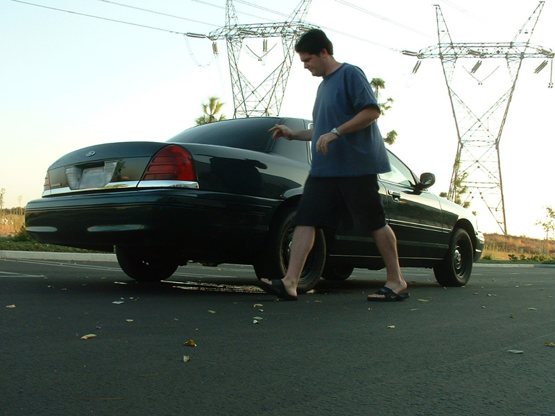 Man standing beside a black Ford sedan on a road under power lines