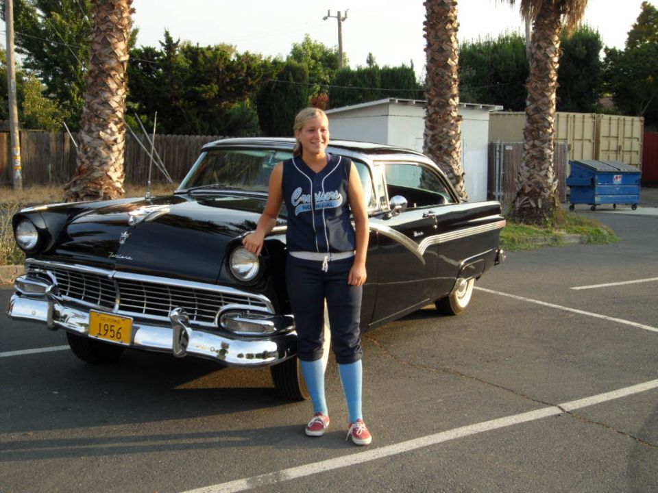 Woman in baseball jersey and pants stands by a black 1956 Ford Fairlane with a yellow license plate