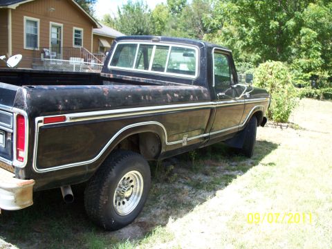 1979 Ford F-250 - Passenger-side rear three-quarter view of a black long-bed Ford F-Series pickup parked on grass beside a tan house, its paint heavily faded…
