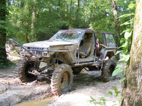 1978 Ford Bronco - A heavily modified Jeep Cherokee XJ navigates a muddy trail through a wooded area.