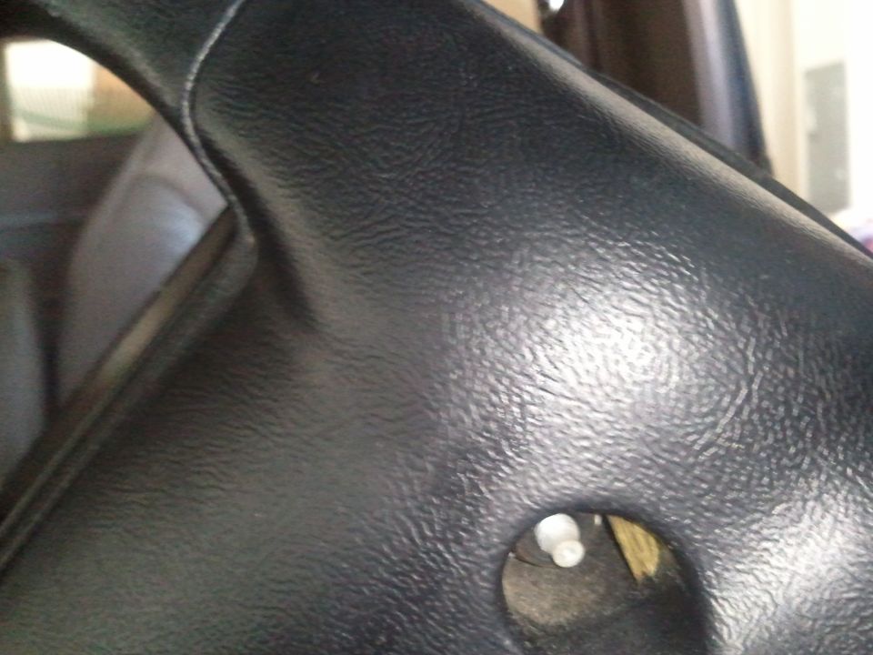 Close-up of a black steering column cover with a hole showing a white plastic piece. Supermotors.net logo visible