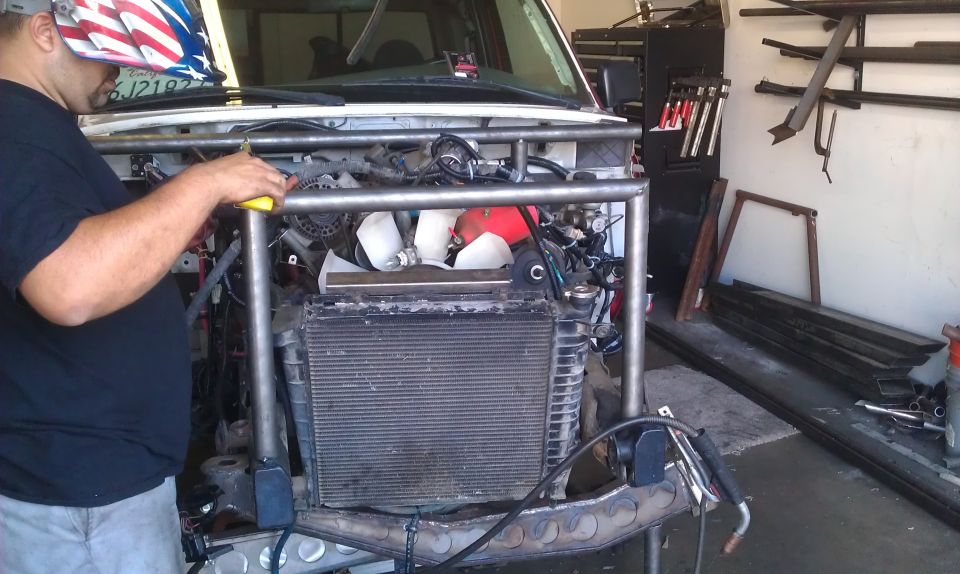 Guy in welding mask working on front end of vehicle with exposed engine and radiator, custom roll cage being installed