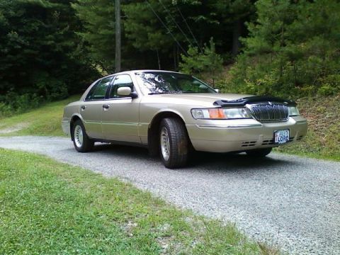 2000 Mercury Grand Marquis - A tan 2000 Mercury Grand Marquis is parked on a gravel road surrounded by trees.