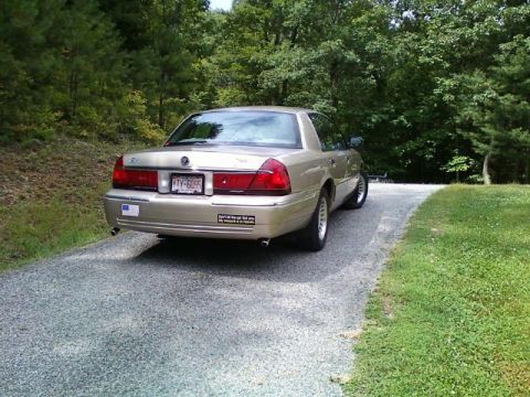2000 Mercury Grand Marquis - A gold 2000 Mercury Grand Marquis sedan is parked on a gravel driveway surrounded by trees.
