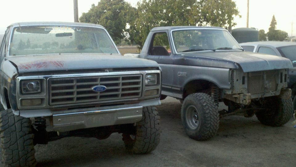 Two lifted Ford trucks parked side-by-side, front view. The truck on the left has a prominent grille and headlights
