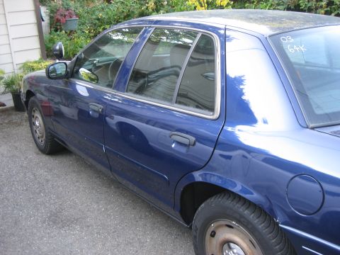 2003 Ford Crown Victoria - A blue 2003 Ford Crown Victoria is seen from the side, parked outdoors with some foliage in the background.
