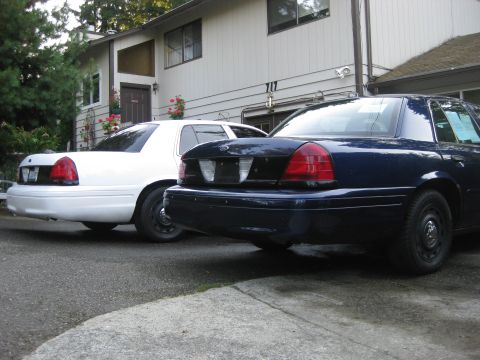 2003 Ford Crown Victoria - Two Ford Crown Victorias, one blue and one white, are parked in front of a house.