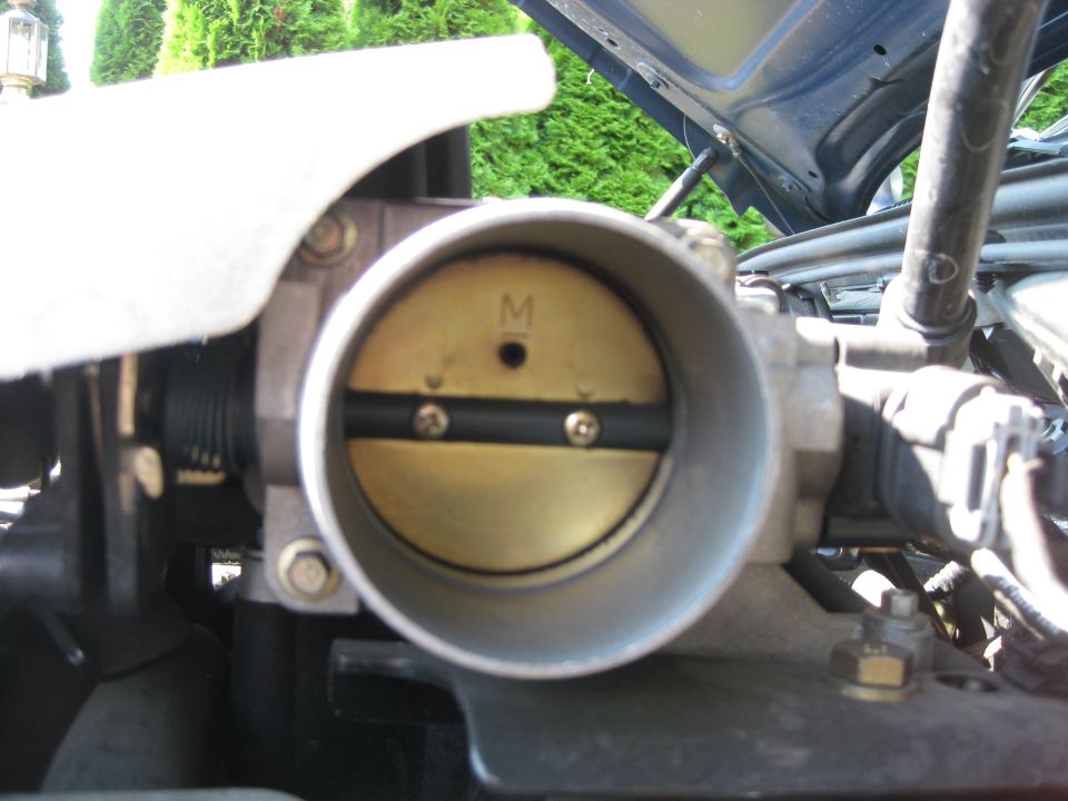 Close-up of a car's throttle body with the butterfly valve visible. There's an 'M' marking on the valve