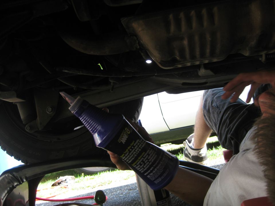 Person performing an oil change under a car, holding a bottle of oil additive
