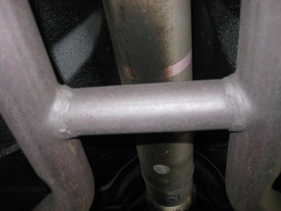 Underside view of a vehicle's exhaust system with a crossbar and drive shaft visible. The metal parts show some wear and dirt