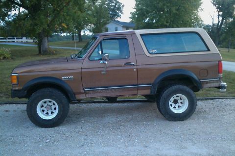 1987 Ford Bronco - A brown 1987 Ford Bronco with a tan top is parked on a gravel surface, viewed from the side.