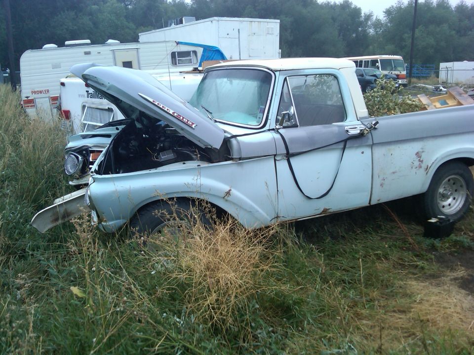 Light blue and gray Ford F-series truck with hood open, parked in tall grass near trailers