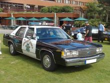 1991 Ford LTD Crown Victoria - This police car, likely a 1991 Ford Crown Victoria, is parked on a grassy lawn in front of a building.